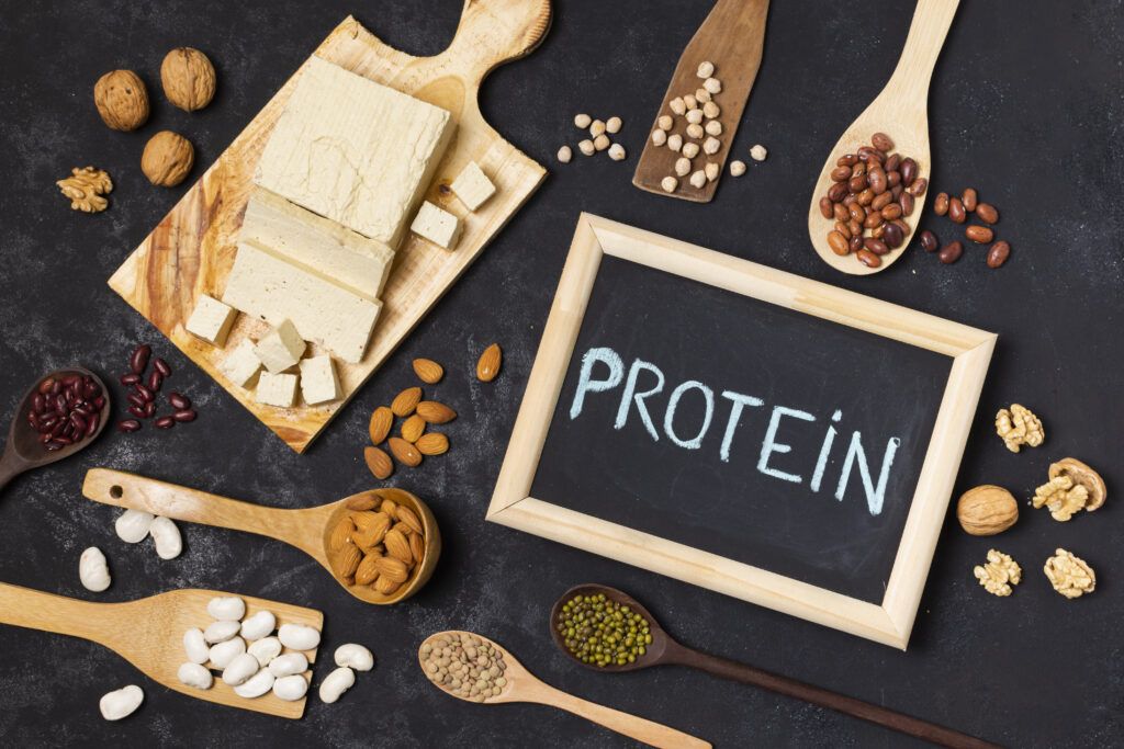 Variedad de alimentos ricos en proteínas, incluyendo legumbres, carnes, frutos secos y huevos, con un cartel destacado en el centro que dice 